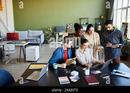 Young multicultural employees gathered by workplace in front of laptop discussing online video and making notes at meeting in office Stock Photo