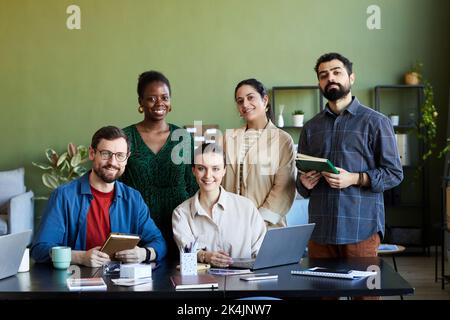 Team of young successful designers gathered by workplace looking at camera with smiles while working over new business project Stock Photo