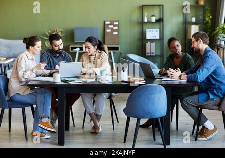 Two small groups of modern creative designers discussing working points while sitting by table in front of laptops and brainstorming Stock Photo