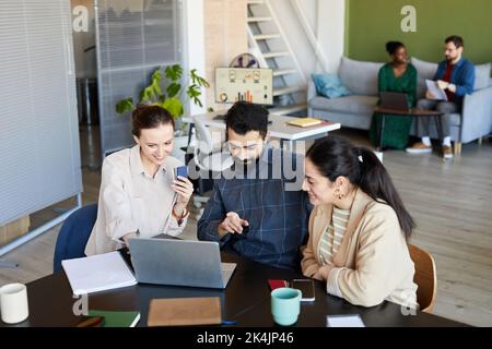 Confident man pointing at laptop screen while discussing data or video with two young female colleagues watching online training Stock Photo