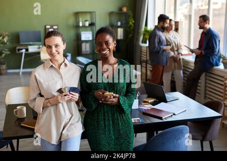 Two young intercultural successful managers or designers looking at camera while standing by their workplace against coworkers Stock Photo