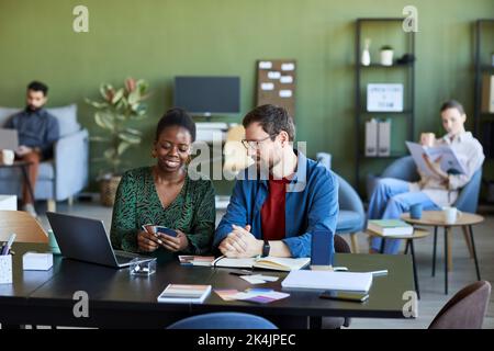 Happy young female designer showing color palette to male colleague whlle both sitting by workplace and preparing new project Stock Photo