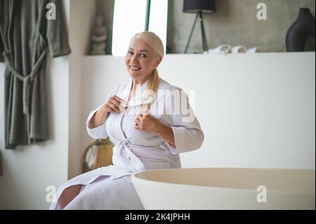 Joyous beautiful lady combing her ponytail with a hairbrush Stock Photo