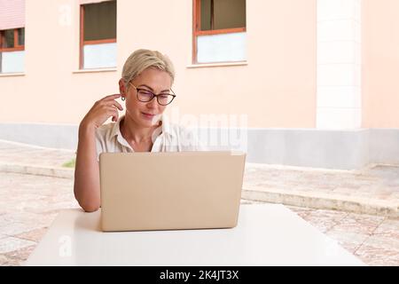 Female freelancer working on laptop at cafe. Portrait of young blond woman sitting alone in cafe outdoors, checking her email while working in her not Stock Photo
