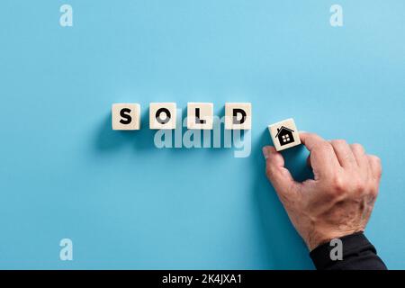Male hand puts the wooden cube with house icon next to the cubes with the word sold. Concept of selling or buying a house. or real estate Stock Photo
