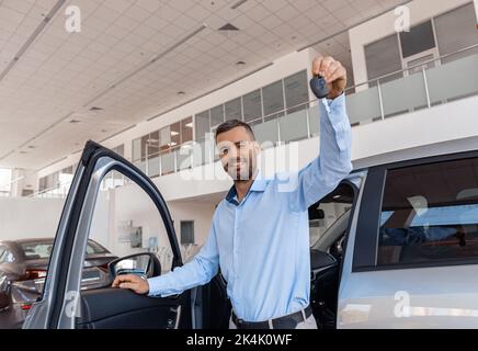 Smiling man customer holding key stands near car in dealership center Stock Photo