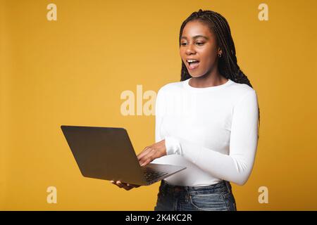 Happy excited African American female student cannot believe her eyes, opened her mouth, standing with open laptop and checking grades, feeling happy from passing the exam, victory concept Stock Photo