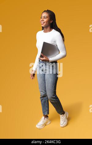 Positive smiling woman with brunette hair standing with closed laptop in hands, while having break. Indoor studio shot isolated on yellow background Stock Photo