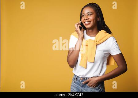 Cheerful smiling woman talking on smartphone and looking away while standing isolated on yellow background. Multiracial lady has pleasant phone conversation Stock Photo