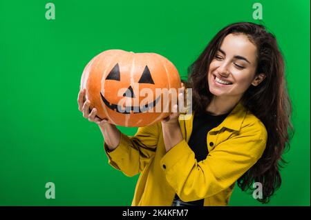 Photo of a happy joyful caucasian woman in casual clothes, holding painted halloween pumpkin, standing on isolated green background, smiling. Halloween party, nightmare concept Stock Photo