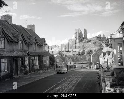 1955, historical, high street, Corfe Castle with the Greyhound Hotel in the distance and the ruins of Corfe Castle on hill-top, Dorset, England, UK. An Austin car of the era is parked on the road, opposite terraced cottages. Dating back to the 11th century, the castle was orginally built by William the Conqueror and was one of the earliest fortifications built partly of stone. In 1645 the medieval castle was slighted, ie partially destroyed after the English Civil War to prevent it being used as a potential fortress and became a ruin.
