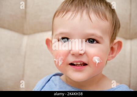 Face cream toddler baby, sofa background in home living room. Close-up portrait of a cute baby with cream on her cheeks. Kid aged one year and two mon Stock Photo