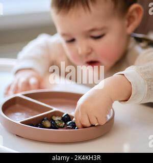 Toddler baby eats fruits and berries with his hand, table close-up. Child hands take food from a beige plate. Kid aged one year and two months Stock Photo