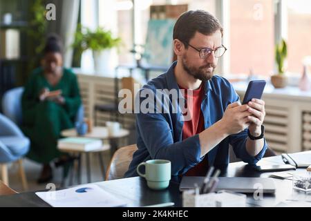 Young serious businessman in eyeglasses and casualwear scrolling in smartphone while looking for online data or watching video Stock Photo