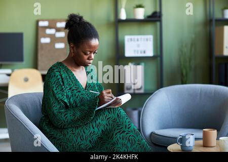 Young serious African American woman in green dress making notes in notepad while sitting in comfortable armchair in office Stock Photo