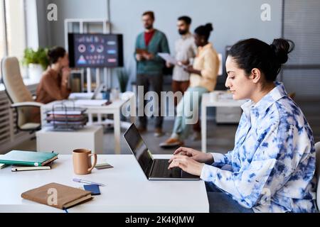 Side view of young serious female employee typing on laptop keypad while sitting by desk in openspace office against her colleagues Stock Photo