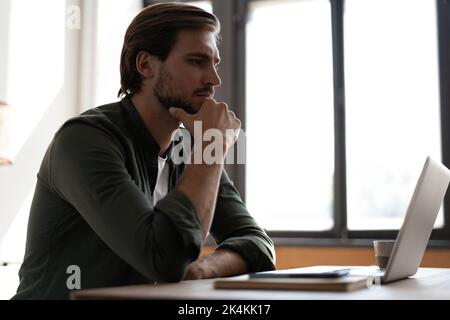 Pensive bearded man sitting at table work at laptop thinking of problem solution, thoughtful male employee pondering considering idea looking at compu Stock Photo