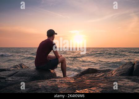 Rear view of man sitting on rock and watching idyllic sunset over ocean. Stock Photo