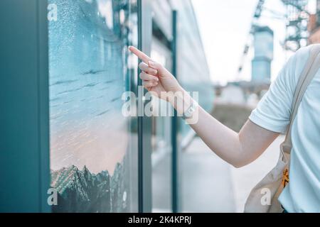 Young tourist woman with backpack stands on city street near information screen on bus stop and chooses route of trip on map. Stock Photo