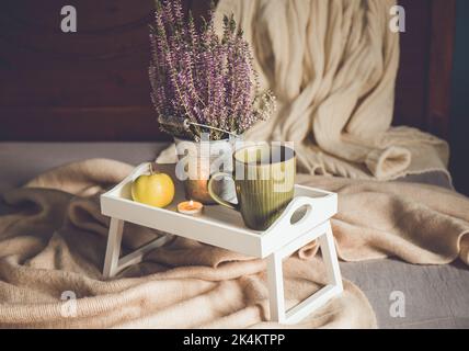 Cute white wooden breakfast tray with legs on home bed with cup of hot tea, tea light candle and heather flower. Autumn arrangement set with scarf. Stock Photo