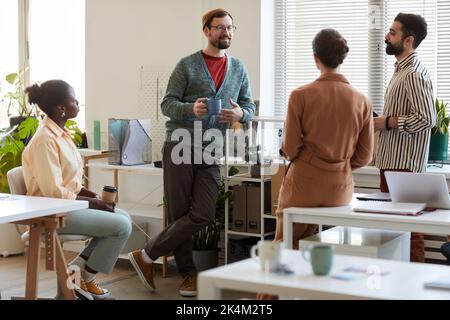 Group of four young intercultural managers discussing latest news or plans for following weekend while having coffee at break Stock Photo