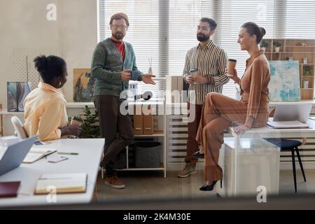 Four young multicultural colleagues with cups of coffee discussing news while businessmen standing in front of two female colleagues Stock Photo