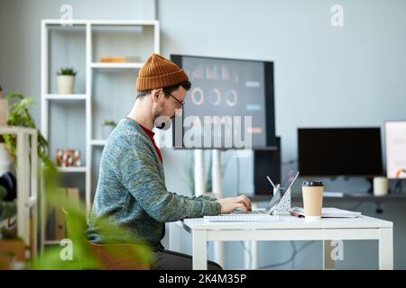 Side view of young serious businessman concentrating on network while sitting by workplace in front of laptop and working over project Stock Photo