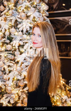Beautiful female with long blonde hair, standing next to Christmas tree, looking at us; she's wearing a black dress Stock Photo