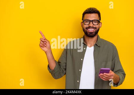 Photo of cute handsome man wear khaki shirt spectacles holding modern device pointing empty space isolated yellow color background Stock Photo