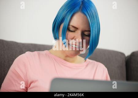 Blue haired woman working on laptop at home. Young white female doing distant work on notebook computer Stock Photo