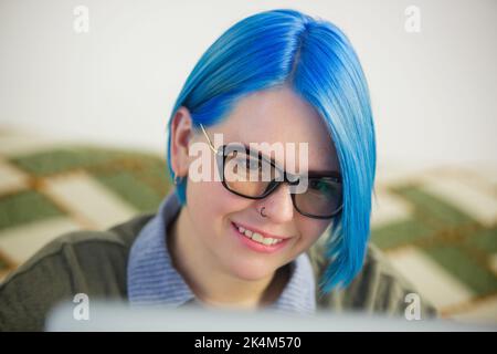 Portrait of happy blue haired woman working on laptop at home. Cheerful white female with dyed hair doing distant work on notebook computer with smile Stock Photo