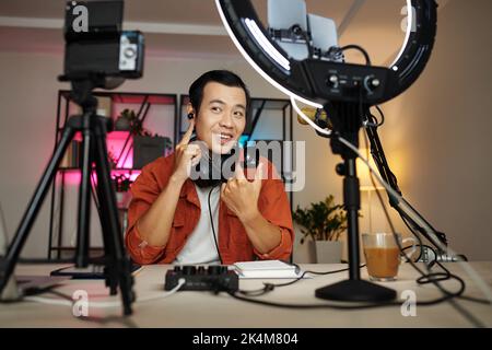 Modern Asian male blogger demonstrating new wireless earphones on camera, making review or advertisment Stock Photo