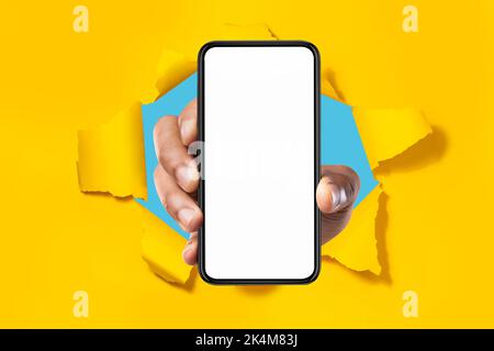 Cellular app. Male hand showing smartphone with white blank screen breaking through torn yellow paper background, mockup Stock Photo