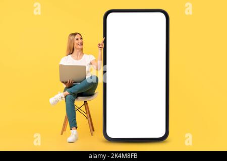 Happy european young woman sit on chair with laptop, pointing finger at big smartphone with blank screen Stock Photo