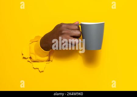 Black male hand holding a mug of coffee through torn yellow paper background, closeup, copy space Stock Photo