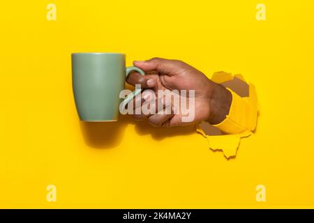 African american man holding cup of hot beverage through torn yellow paper background, closeup, copy space Stock Photo
