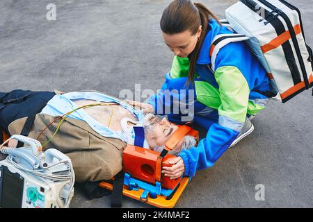 Female paramedic fixing head of male victim with neck injury lying on ambulance stretcher. First aid with cervical collar and defibrillator Stock Photo