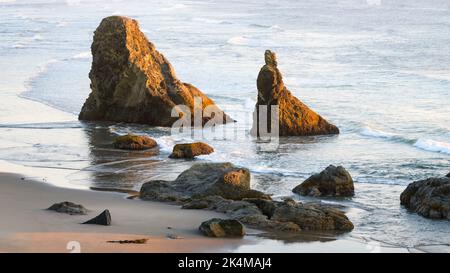 Rocks on the Pacific Ocean beach in Bandon Oregon as the summer sun sets and waves surround the small sea stacks Stock Photo