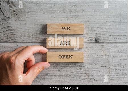 Male hand assembling a We are open sign written on three stacked wooden blocks placed over white wooden background. Stock Photo