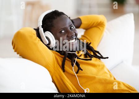 Cheerful Black Guy With Dreadlocks Wearing Headphones Relaxing On Comfortable Couch Stock Photo