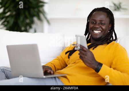 Portrait Of Smiling Black Guy Using Laptop And Reading Message On Smartphone Stock Photo