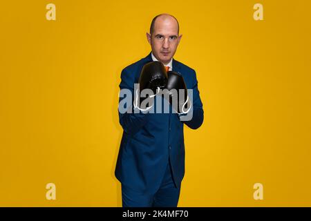 A bald Caucasian adult male wearing a blue suit, white shirt and orange tie is wearing boxing gloves and is in a defensive stance. The background is y Stock Photo