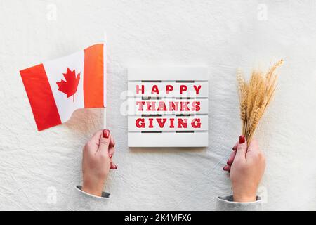 Happy Canada Thanksgiving Day. Canadian flag and wheat ears in hand on off white beige textile. Greeting caption on wood palette. Flat lay, top view. Stock Photo