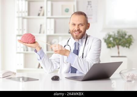 Young doctor neurologist sitting in an office and pointing at a human brain model with laptop computer on the desk Stock Photo