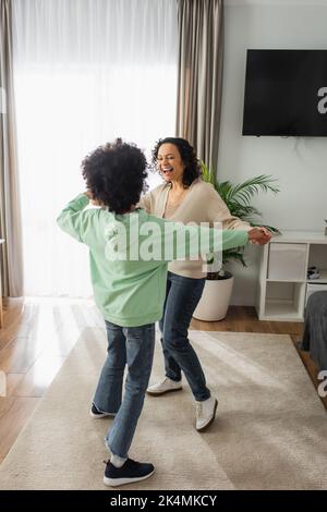 excited african american woman holding hands with curly preteen daughter while dancing at home,stock image Stock Photo