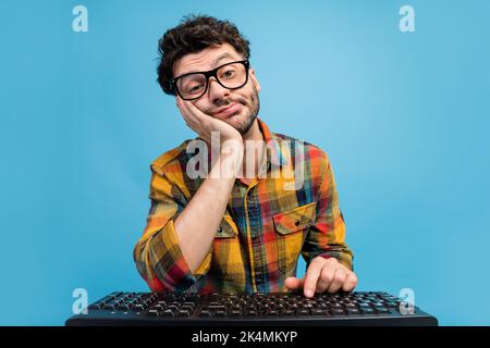 Photo of unhappy tired man in eyeglasses dressed checkered shirt writing message on keyboard hand cheek isolated on blue color background Stock Photo