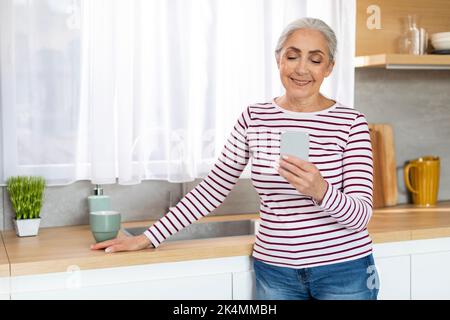 Portrait Of Beautiful Smiling Elderly Woman Resting With Smartphone In Kitchen Interior Stock Photo