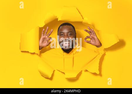 Everything is under control. Happy black man giving positive opinion, making ok gesture posing in torn yellow paper hole Stock Photo