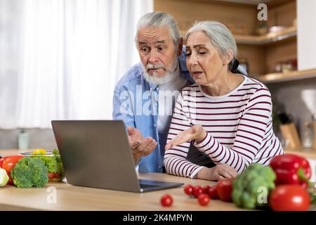 Confused Senior Spouses Suffering Problems With Laptop While Spending Time In Kitchen Stock Photo