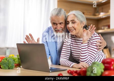 Cheerful Elderly Spouses Making Video Call With Laptop In Kitchen Interior Stock Photo